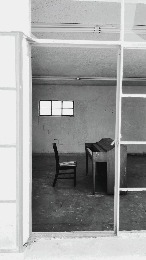 black and white photo through a paneless window of a lone chair in front of broken down piano