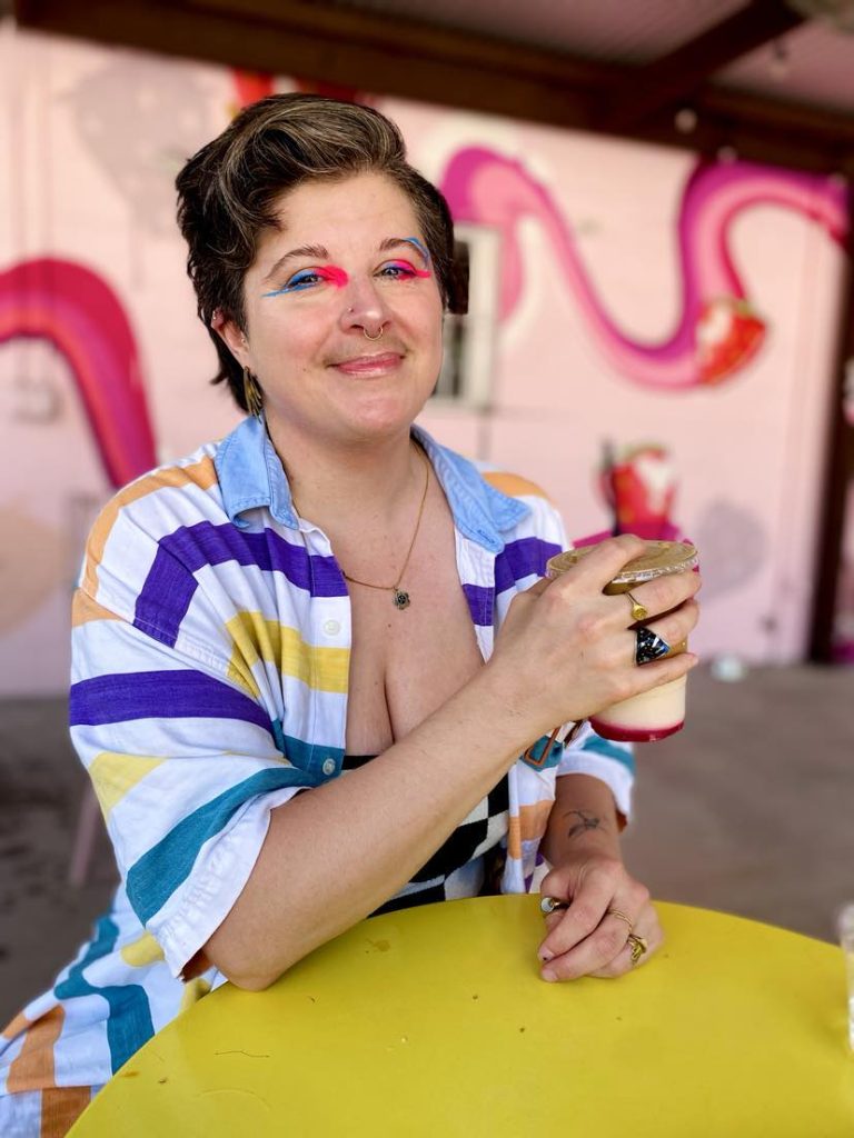 nonbinary person with bright makeup holding a coffee and smiling